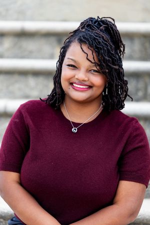 Smiling young Black woman with braided locs updo, wearing burgundy sweater and silver necklace, seated on stone steps.