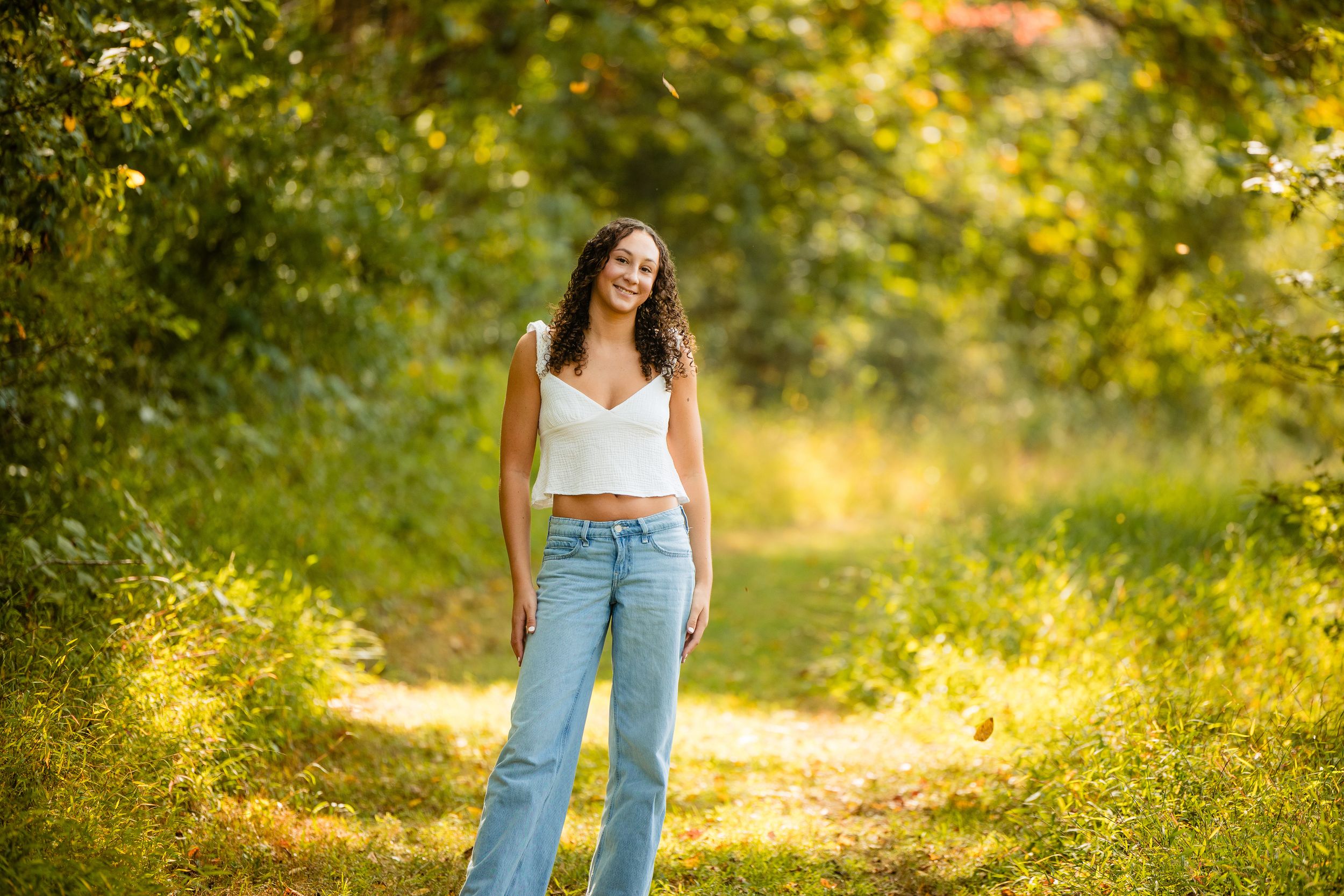 Young woman in white crop top and blue jeans smiling on a sunny forest path surrounded by lush green trees.