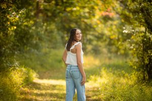 Young woman in white top and jeans smiling over shoulder on sunlit forest path.