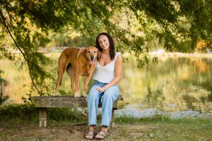 Young woman sitting on bench with golden retriever dog by a peaceful lake surrounded by green trees.