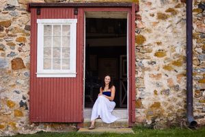 A smiling woman in a white skirt sits in the doorway of a rustic stone building with a red wooden door.