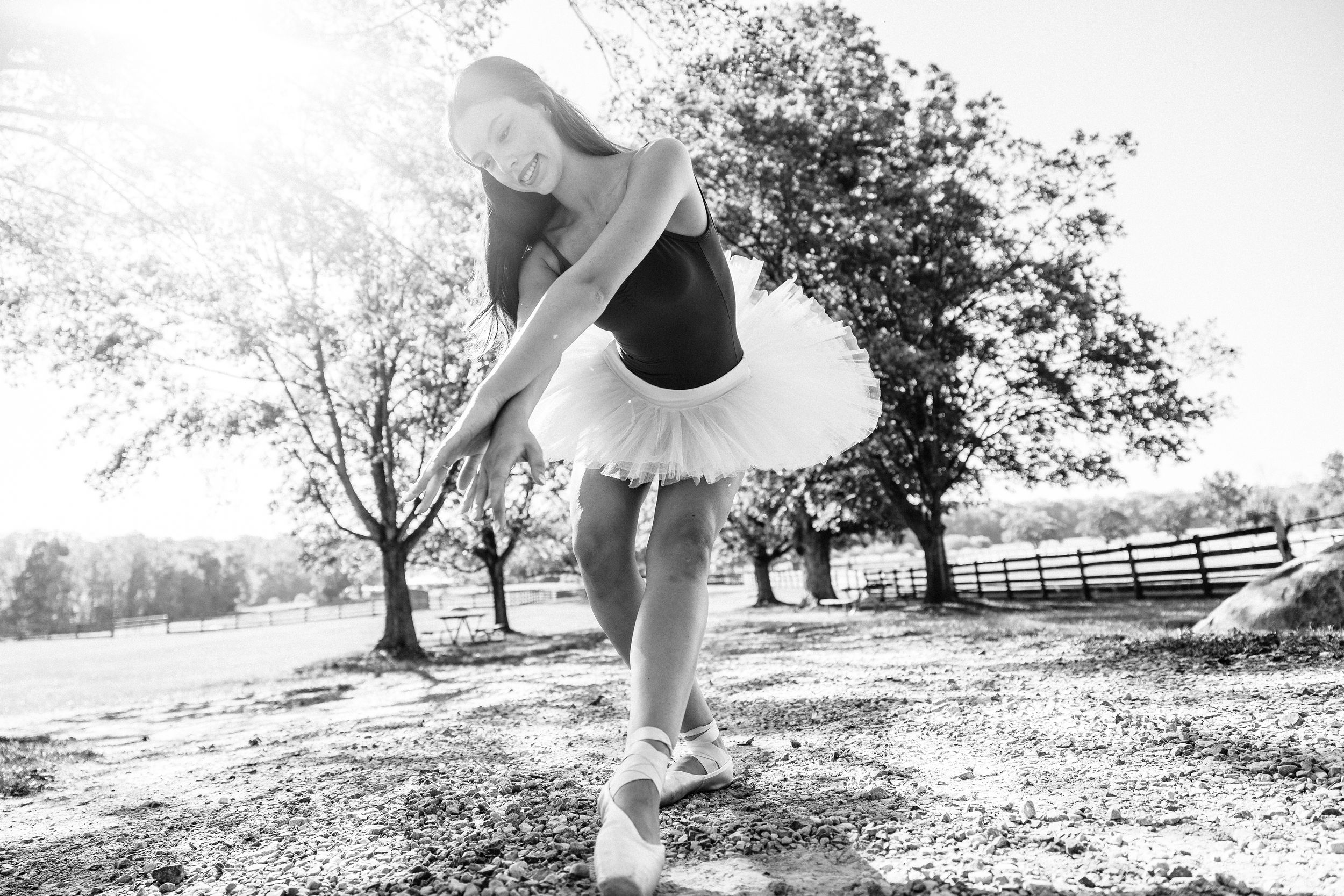Black and white photo of a ballerina in tutu performing graceful pose outdoors among trees in natural light.
