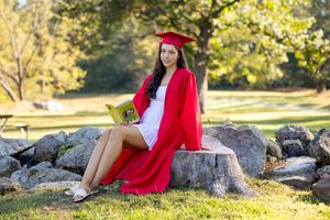 Young woman in red graduation cap and gown sitting on rocks reading a book outdoors.