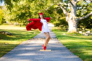 Graduate in white dress and red cap and gown twirls joyfully on a park path surrounded by green trees.