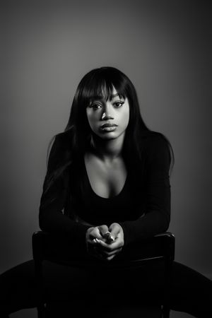 Black and white portrait of a young woman in black top sitting confidently with hands clasped in studio setting.