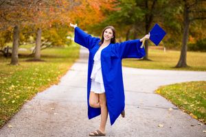 A joyful graduate in a blue cap and gown celebrates outdoors on a path surrounded by colorful autumn trees.