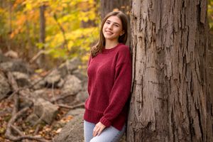 Young woman in red sweater smiling while leaning against a tree in an autumn forest setting.
