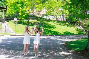 Two young women in white dresses play with hula hoops outdoors on a sunny day surrounded by lush green trees.