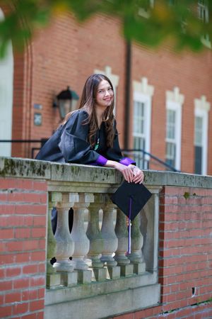 Young woman in graduation gown and cap leaning on stone balustrade at university campus, smiling proudly.
