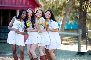 Four women in white dresses holding yellow sunflowers pose together outdoors near a red barn.