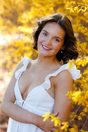 Smiling young woman in white ruffled dress posing outdoors among vibrant yellow autumn foliage.