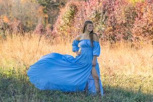 Young woman in flowing blue off-shoulder gown poses gracefully in golden autumn field with colorful foliage backdrop.