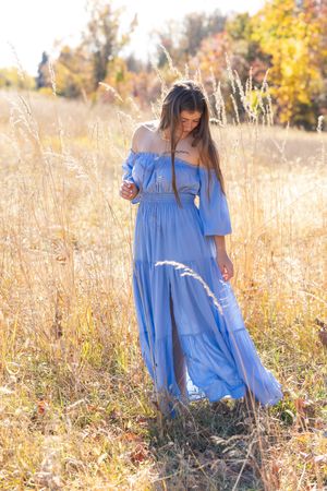 Woman in blue off-shoulder maxi dress standing in golden field during autumn, sunlight illuminating the scene.