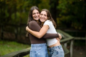 Two smiling young women hugging each other outdoors near a wooden fence with green trees in the background.