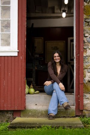 Smiling young woman with dark hair sitting on stone steps of a red barn doorway wearing jeans and brown top.