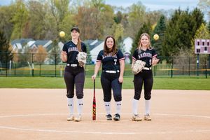 Three softball players in black uniforms pose together on a field with red dirt and residential houses in background.