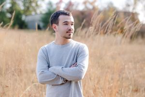 Young man in gray sweatshirt standing in golden field with arms crossed, looking away thoughtfully.