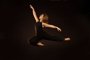 A dancer in black attire leaps gracefully against a dark background, arms extended and legs split mid-air.