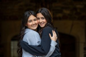 Two young girls with dark hair smiling and hugging each other outdoors near a stone building.