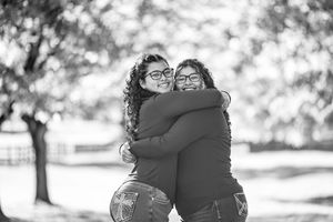 Two young women with curly hair and glasses share a warm hug outdoors in a black and white photo.