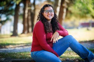 Young woman with curly hair and glasses smiling outdoors, wearing a red top and jeans, seated in a sunny park.