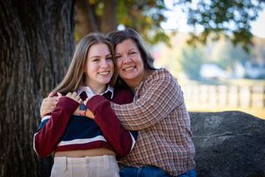Two women smiling and hugging outdoors near a large tree, sharing a warm embrace in a fall setting.