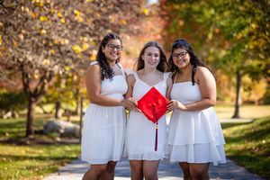 Three people in matching white dresses and coordinated outfits pose together outdoors during autumn.