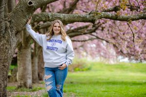 Person in white sweatshirt and jeans poses beneath blooming pink cherry blossom trees in park setting.