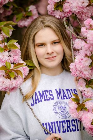 Student wearing James Madison University sweatshirt poses among blooming pink cherry blossoms in spring.