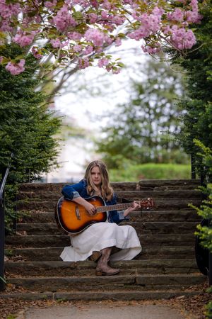 A musician sits on stone garden steps beneath blooming cherry blossoms playing an acoustic guitar on a spring day.