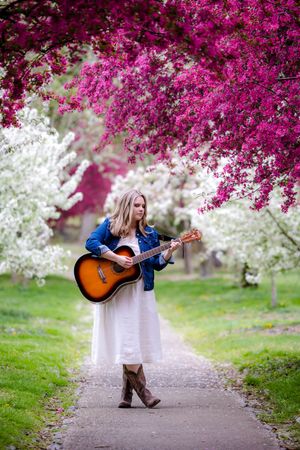 A guitarist in a white dress and cowboy boots plays under vibrant pink cherry blossoms on a spring path.