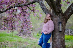 Springtime portrait under blooming cherry blossoms with someone in a pink sweater and blue jeans leaning against a tree.
