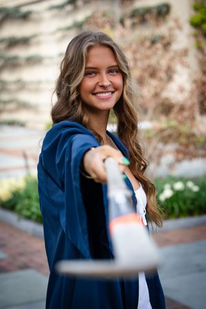 A person in a navy blue graduation gown stands outside smiling on a brick pathway near flowering plants.