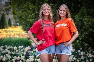 Two people in bright orange and red shirts pose together in a garden filled with blooming flowers.