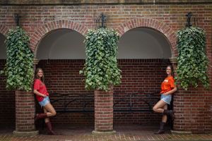 Two young women in orange shirts and boots pose against a brick wall with ivy-covered arches and columns.
