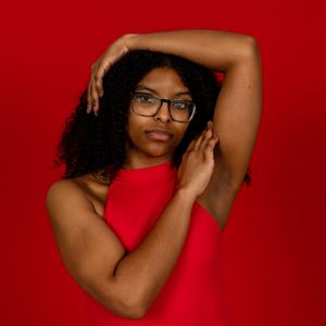 Portrait against red background features person in matching red sleeveless top, black glasses and natural curly hair.