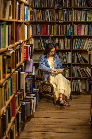 Someone reads quietly in wooden chair surrounded by tall bookshelves in cozy library corner.