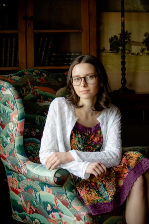 Portrait on a floral upholstered chair showing white cardigan, patterned dress and glasses in a cozy home setting.