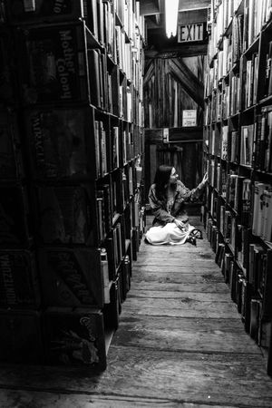 A reader sits between towering bookshelves in a dimly lit library with wooden floors and an exit sign visible in the distance.