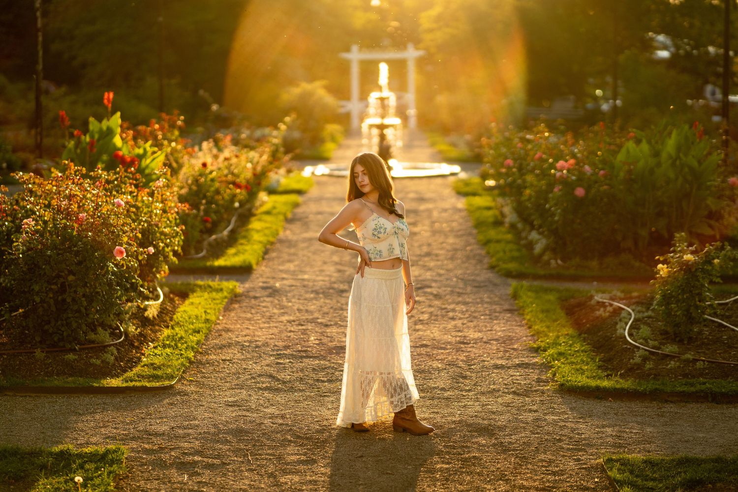 Athlete in black tank top and white pants sits on a paved path surrounded by lush green trees during outdoor workout.