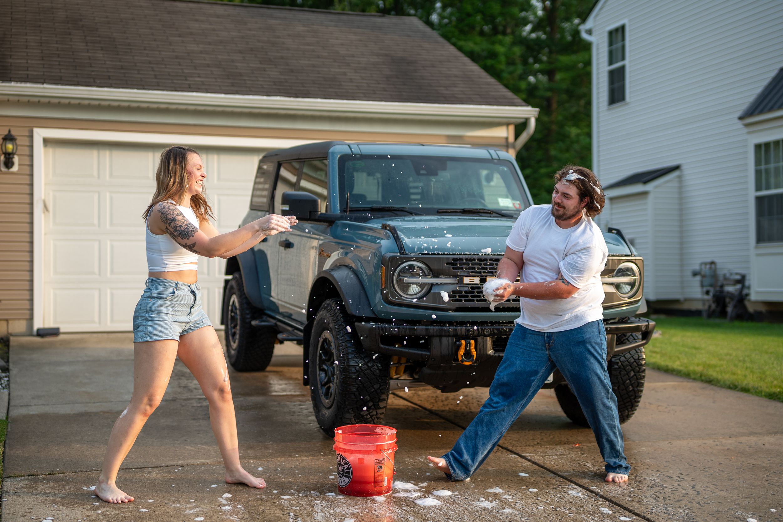 Two people playfully splash each other with sponges while washing a lifted black Bronco in a residential driveway.