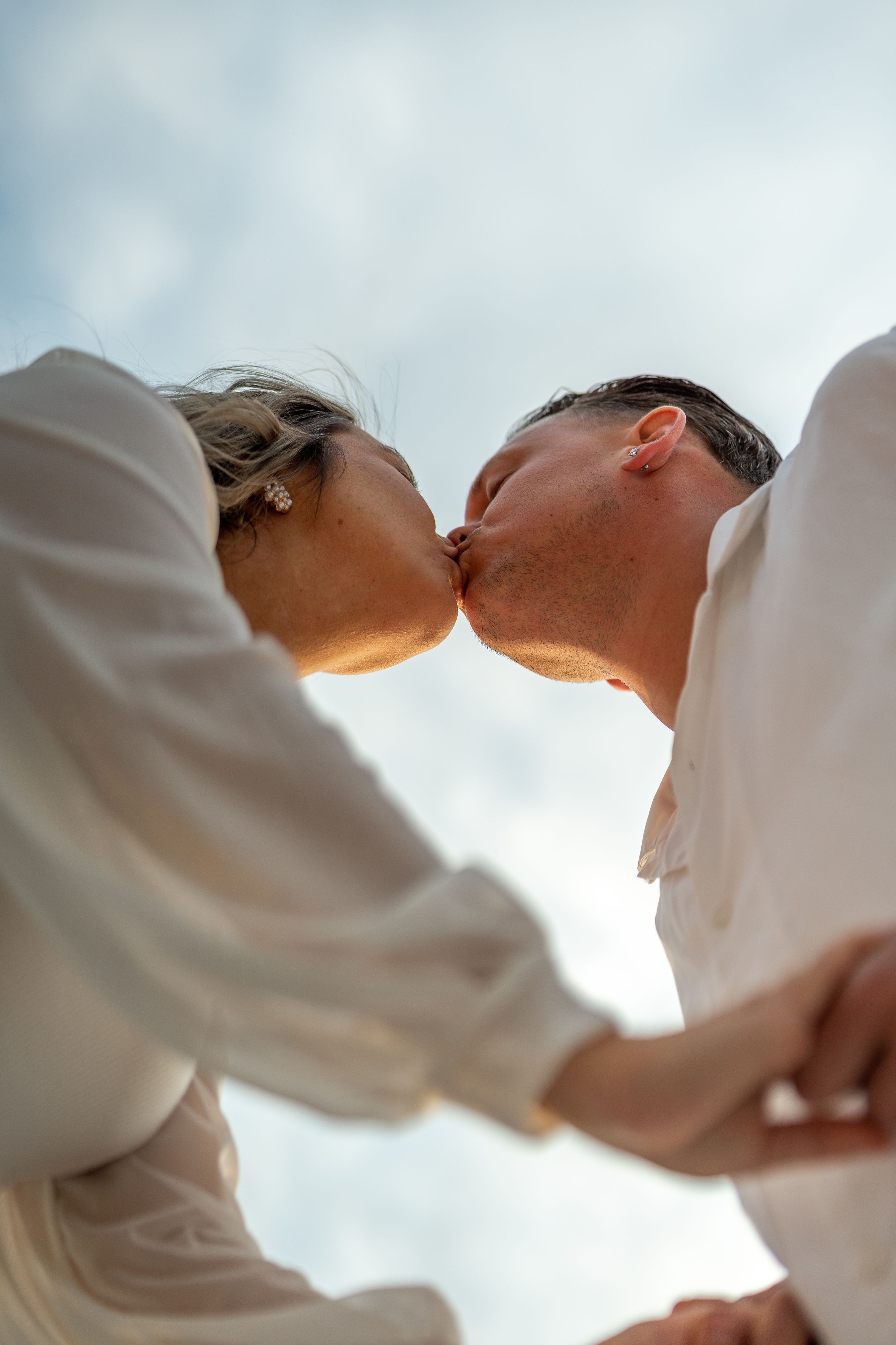Intimate couple's kiss silhouetted against cloudy sky in white clothing.