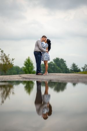 Romantic couple embracing by still water with perfect reflection under cloudy sky.