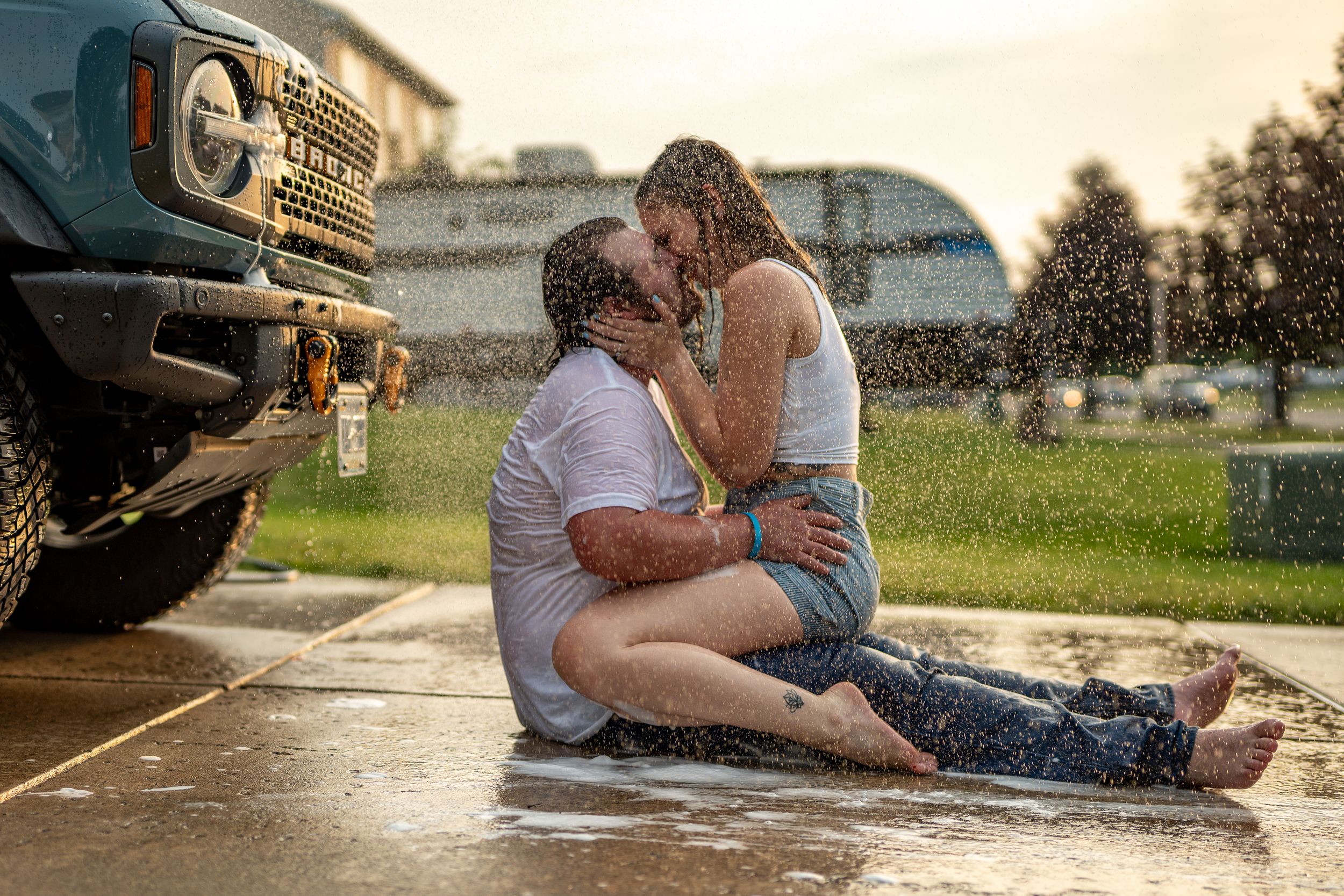 Romantic couple embraces in the rain next to a pickup truck at sunset in a grassy field.