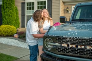 Couple laughs while washing muddy Bronco truck in driveway.