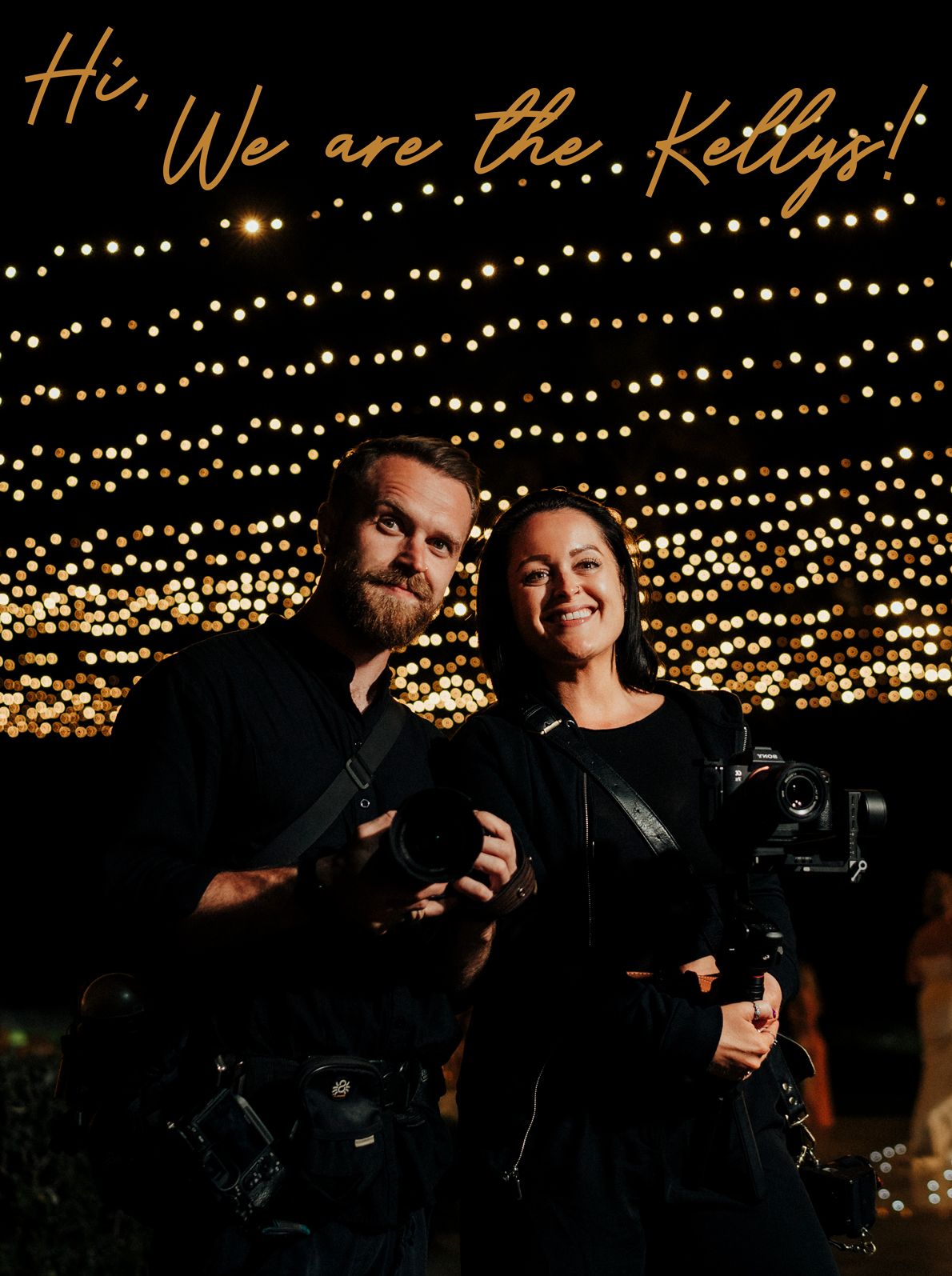 Two photographers smile under twinkle lights at night holding professional cameras.