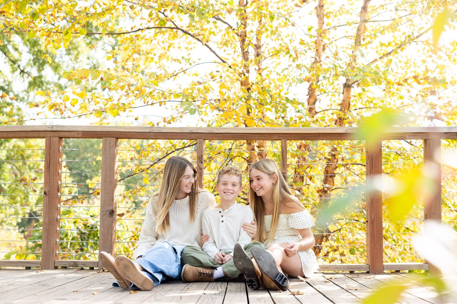 A group of three young women sitting together on a wooden deck, surrounded by autumn foliage.