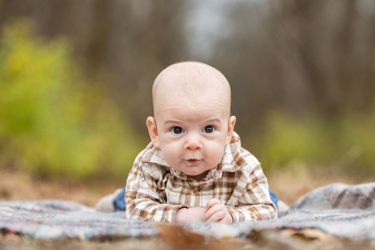 A smiling baby lying on a blanket in a natural outdoor setting, surrounded by autumn foliage.