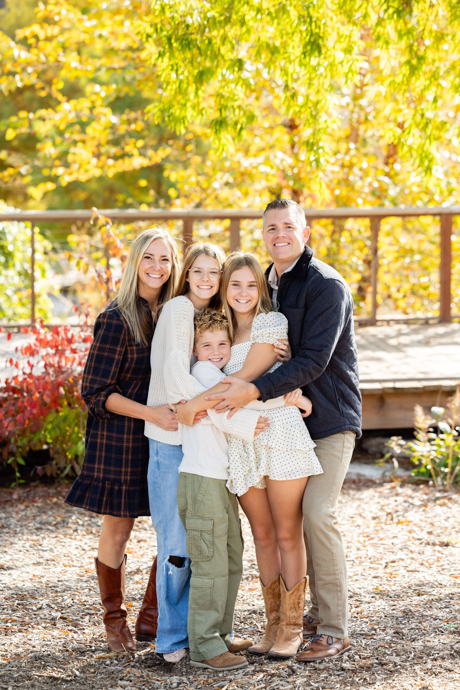 A family of four, including two adults and two children, stand together in a park or garden setting.