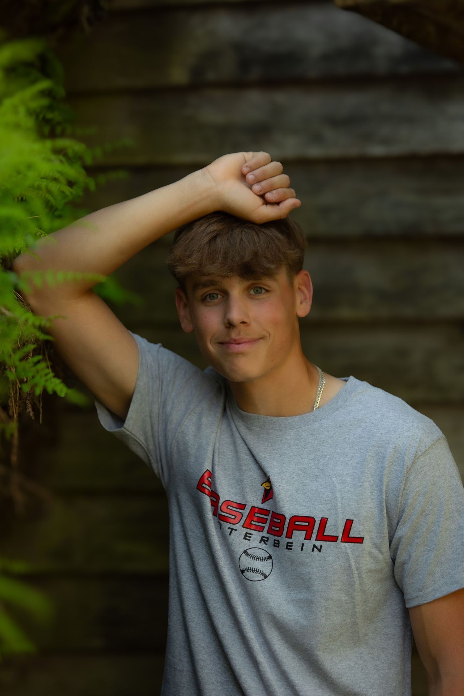 Person wearing a grey baseball t-shirt poses against wooden wall with green foliage in natural lighting.